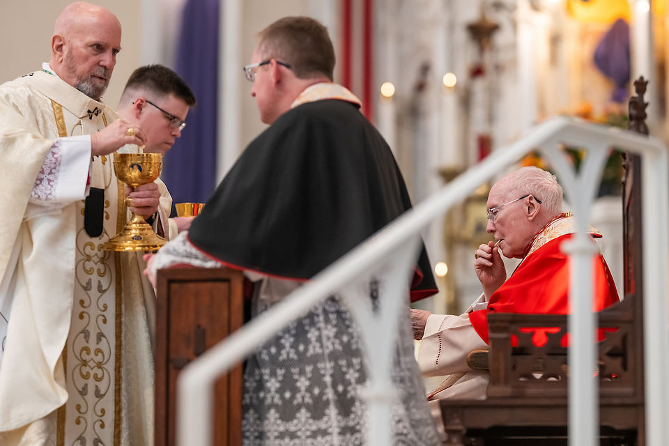 Clergy in ornate robes participate in a church ceremony. One holds a chalice, another in red sits solemnly, with candles in the background.