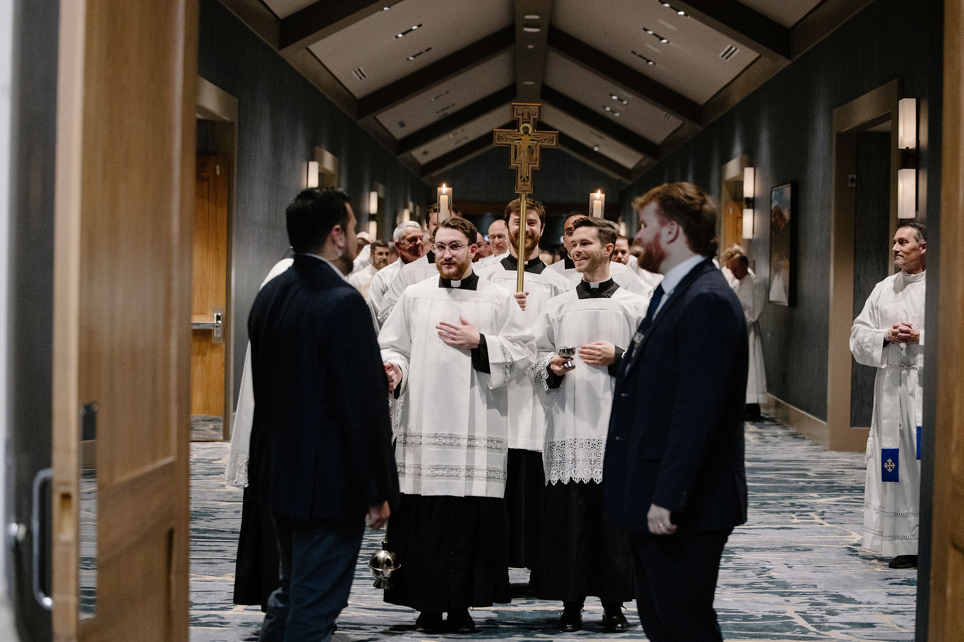Catholic clergy in white vestments walk in procession down a hallway, carrying candles and a crucifix, as attendees stand on either side greeting them.