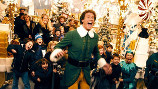 Elf-costumed man joyfully leads excited crowd in festive, decorated store with Christmas trees, lights, and gifts; cheerful atmosphere.