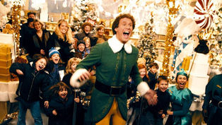 Elf-costumed man joyfully leads excited crowd in festive, decorated store with Christmas trees, lights, and gifts; cheerful atmosphere.