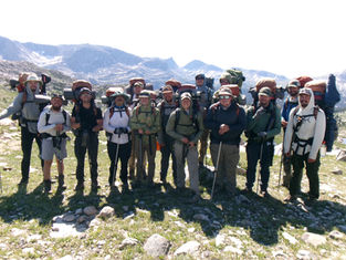 Smiling backpackers pose with trekking poles and loaded packs on a rocky alpine trail, mountains and snowfields behind them.