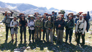 Smiling backpackers pose with trekking poles and loaded packs on a rocky alpine trail, mountains and snowfields behind them.