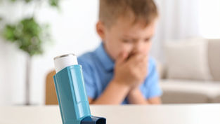 A blue asthma inhaler on a table; blurred background shows a child in blue, hands covering mouth, in a bright room with a plant.