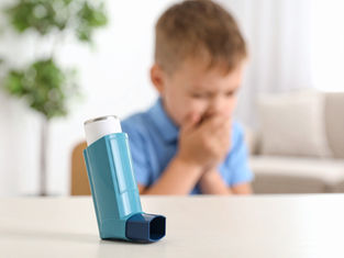 A blue asthma inhaler on a table; blurred background shows a child in blue, hands covering mouth, in a bright room with a plant.