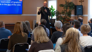 Priest speaks to seated audience in a church meeting room beside a blue slide about Ignatius; crucifix and plants in back.