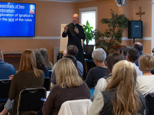 Priest speaks to seated audience in a church meeting room beside a blue slide about Ignatius; crucifix and plants in back.