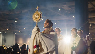 A man holds a golden monstrance in a dimly lit church, surrounded by clergy in white robes, with a spotlight creating a solemn mood.