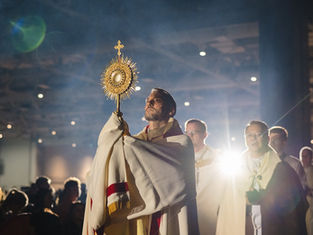 A man holds a golden monstrance in a dimly lit church, surrounded by clergy in white robes, with a spotlight creating a solemn mood.