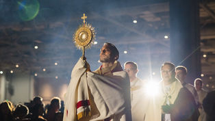 A man holds a golden monstrance in a dimly lit church, surrounded by clergy in white robes, with a spotlight creating a solemn mood.