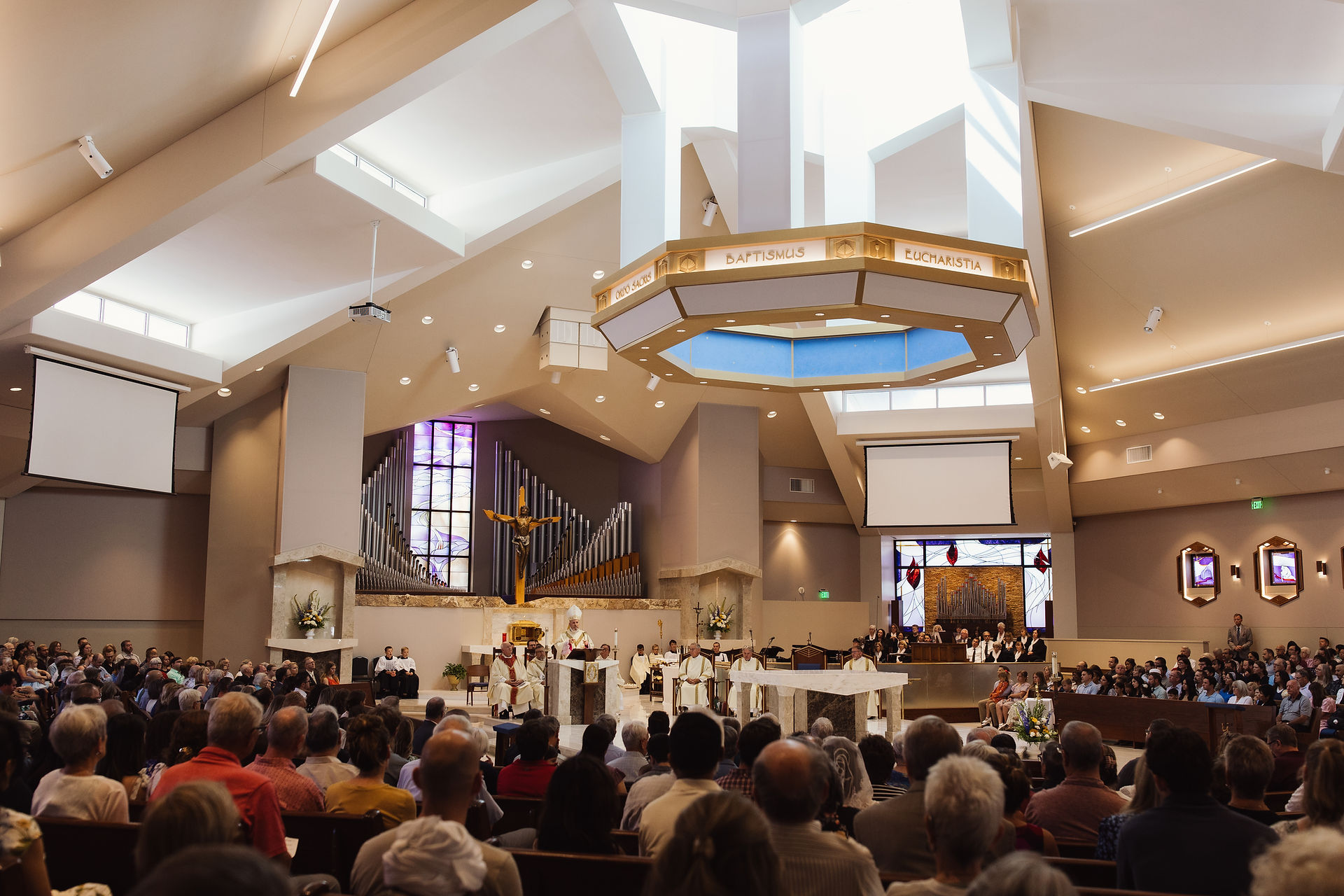 Church service in progress with a priest speaking at the altar. Stained glass and organ pipes in the background, large crowd seated.