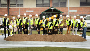 People in hard hats and yellow vests participate in a groundbreaking ceremony, holding shovels over soil, with a truck and building in the background.