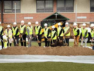 People in hard hats and yellow vests participate in a groundbreaking ceremony, holding shovels over soil, with a truck and building in the background.