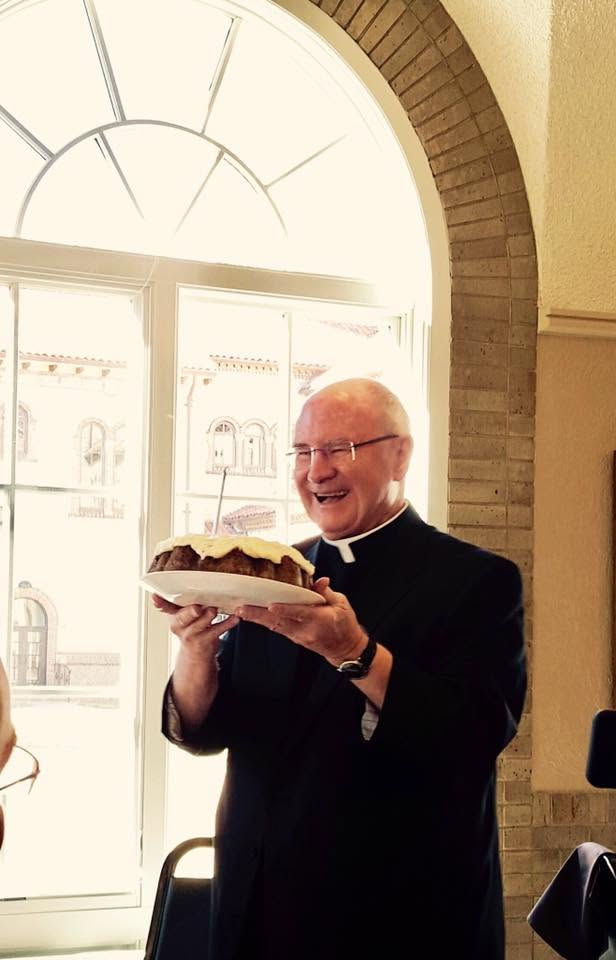 Smiling priest in black cassock holds a cake on a plate beside an arched window in a bright room.