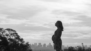 Pregnant woman in profile stands outdoors, city skyline in background. Overcast sky, serene mood, silhouette effect.