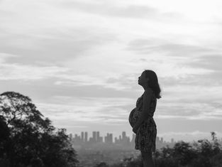 Pregnant woman in profile stands outdoors, city skyline in background. Overcast sky, serene mood, silhouette effect.