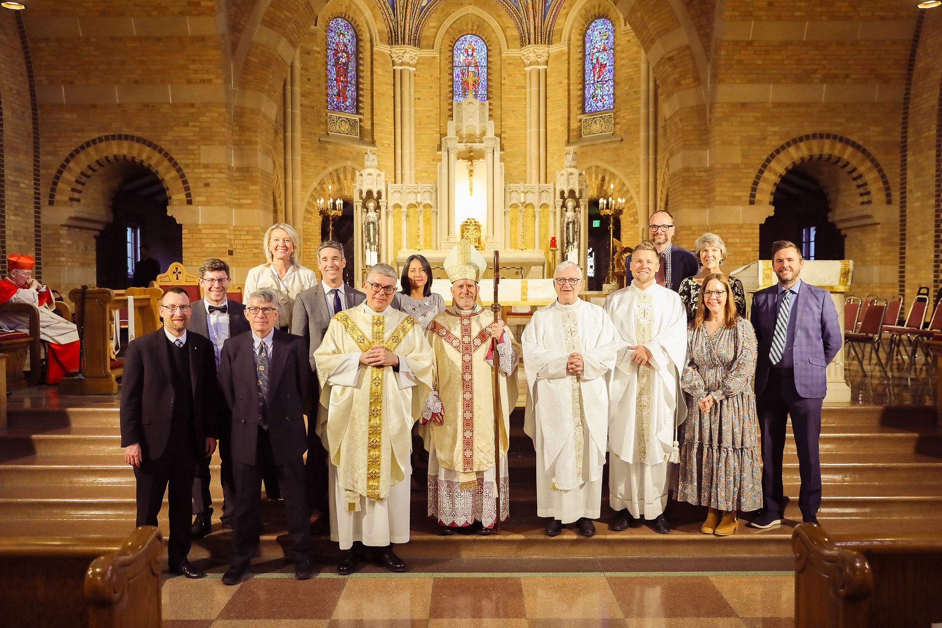 A group of men and women stand with an archbishop in an ornate brick chapel following a religious ceremony. The mood is celebratory.