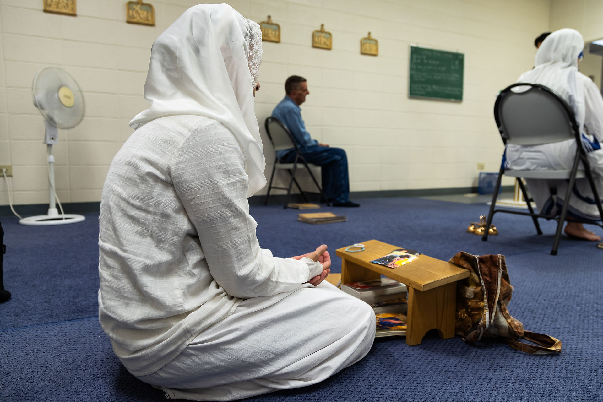Person in white attire kneeling on blue carpet, meditating near a wooden stand with books. A seated person in blue is in the background.