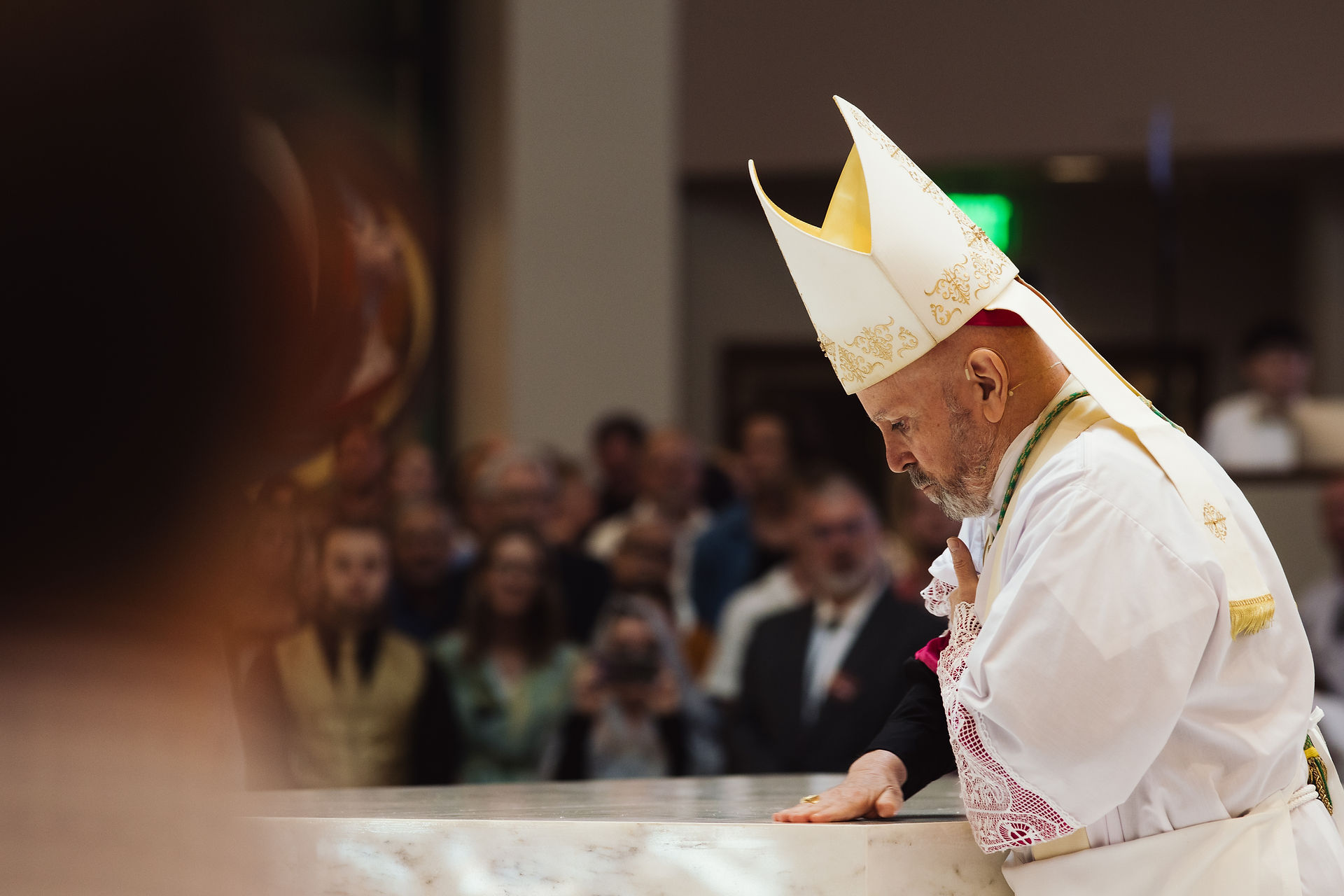 A bishop in a white robe and mitre bows solemnly at an altar. The background is filled with blurred people, suggesting a church setting.