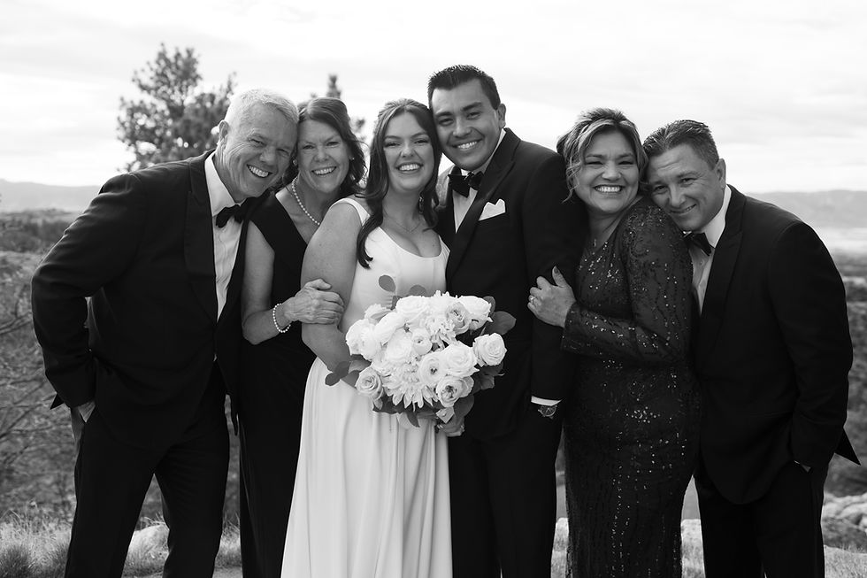 Wedding group photo with a smiling couple in formal attire, surrounded by happy family. Bride holds white roses. Outdoor scenic backdrop.
