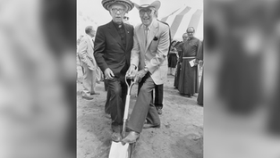 Two smiling men in hats pose on a shovel at a groundbreaking ceremony, with tents and clergy in the background.
