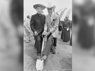 Two smiling men in hats pose on a shovel at a groundbreaking ceremony, with tents and clergy in the background.
