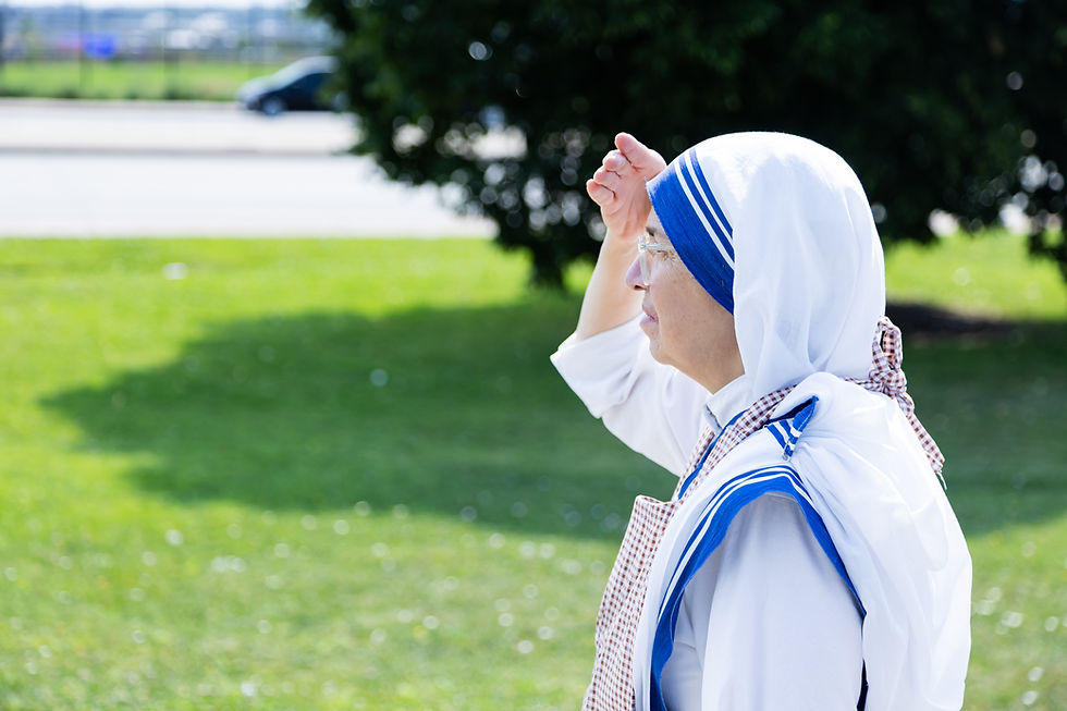 A person in a white and blue habit raises their hand to shield their eyes from the sun. They stand on a grassy area beside a road.