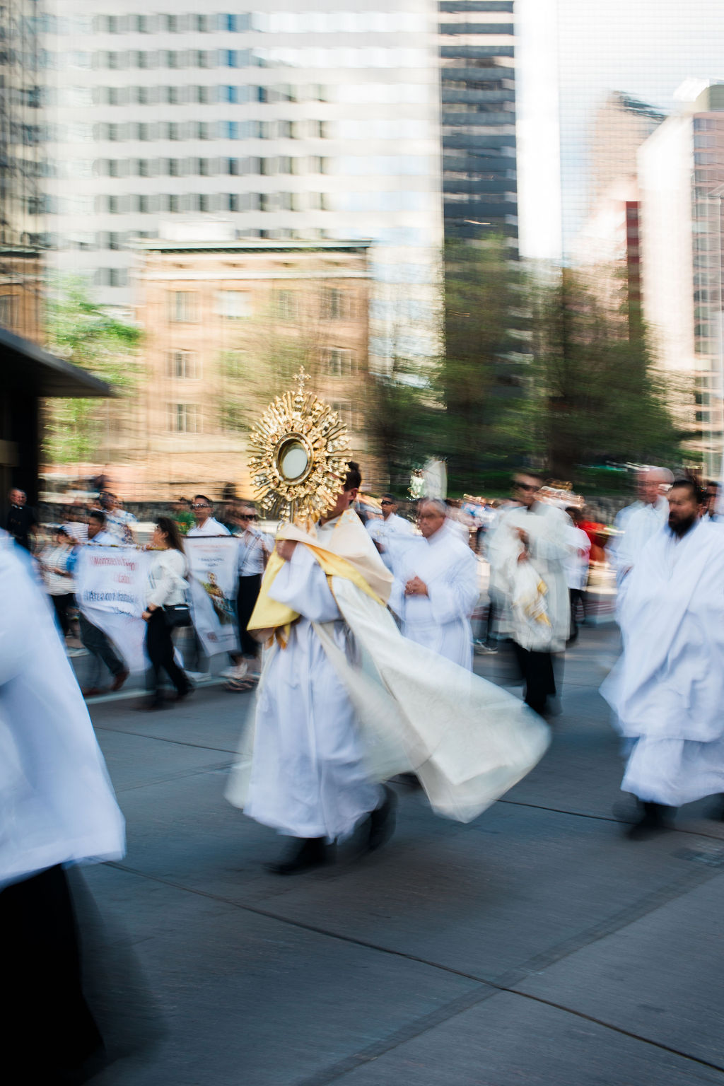 Priests in white robes carry a golden monstrance in a blurred city street procession, with spectators and tall buildings.