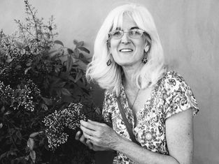 Smiling white-haired woman in glasses holds flowering branches beside leafy plants against a plain wall.