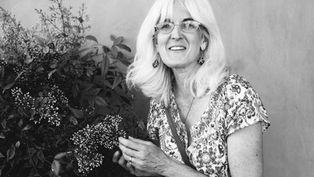 Smiling white-haired woman in glasses holds flowering branches beside leafy plants against a plain wall.