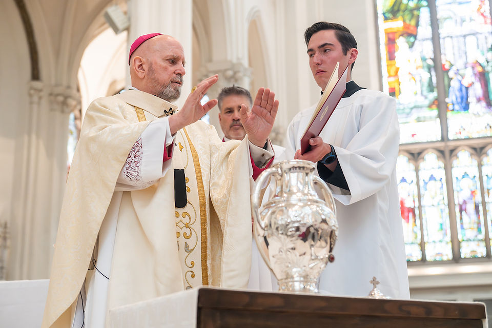 Bishop in ornate robe blesses silver urn in church, assisted by a young man holding a book. Stained glass windows in the background.
