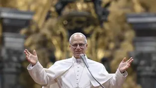 A man in white religious robes speaks into a microphone with arms raised. Ornate golden background. Calm and welcoming expression.