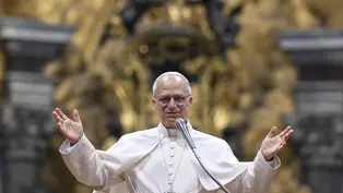 A man in white religious robes speaks into a microphone with arms raised. Ornate golden background. Calm and welcoming expression.