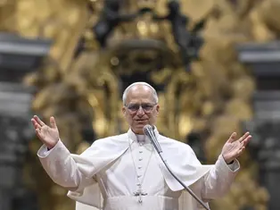 A man in white religious robes speaks into a microphone with arms raised. Ornate golden background. Calm and welcoming expression.