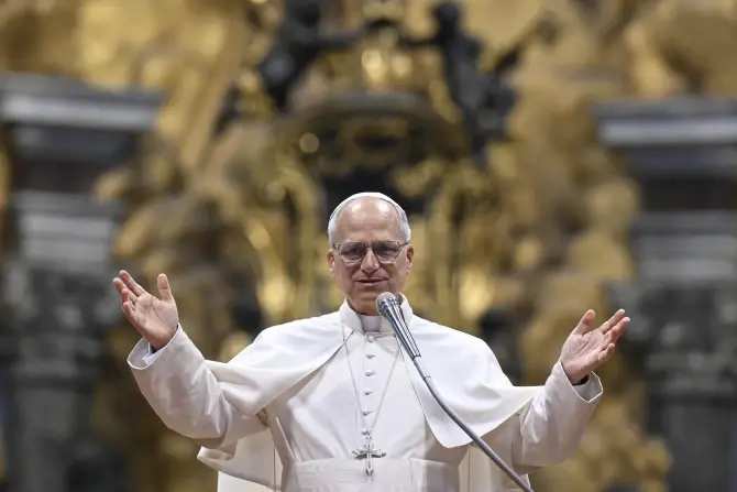 A man in white religious robes speaks into a microphone with arms raised. Ornate golden background. Calm and welcoming expression.