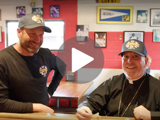 Two men in matching hats and shirts smile in a restaurant with red and white walls, decorated with framed photos. A play button is overlaid.