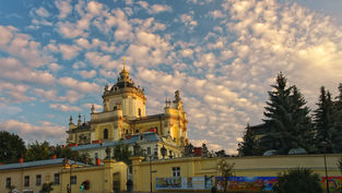 Baroque-style building under a sky with scattered clouds. Trees and a colorful mural adorn the foreground, creating a serene atmosphere.