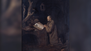 A monk kneels, praying at a desk with an open book in a dark, rustic room. A tree branch and animal skull create a somber mood.