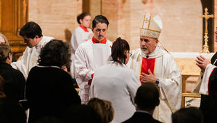 Priest in ornate robe blesses congregation inside a church. Attendees in dark clothing. Background shows altar and gold cross.