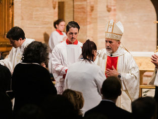 Priest in ornate robe blesses congregation inside a church. Attendees in dark clothing. Background shows altar and gold cross.