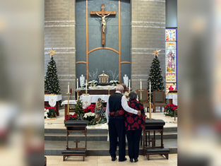 Older couple embracing in a church, facing an altar with Christmas trees and poinsettias. Stained glass and crucifix in the background.