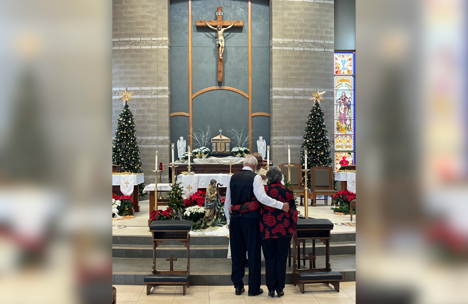 Older couple embracing in a church, facing an altar with Christmas trees and poinsettias. Stained glass and crucifix in the background.