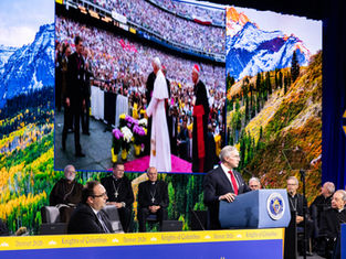 Man speaks at a Knights of Columbus Denver 2026 podium, with bishops seated behind and mountain scenes on giant screens.