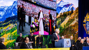 Man speaks at a Knights of Columbus Denver 2026 podium, with bishops seated behind and mountain scenes on giant screens.