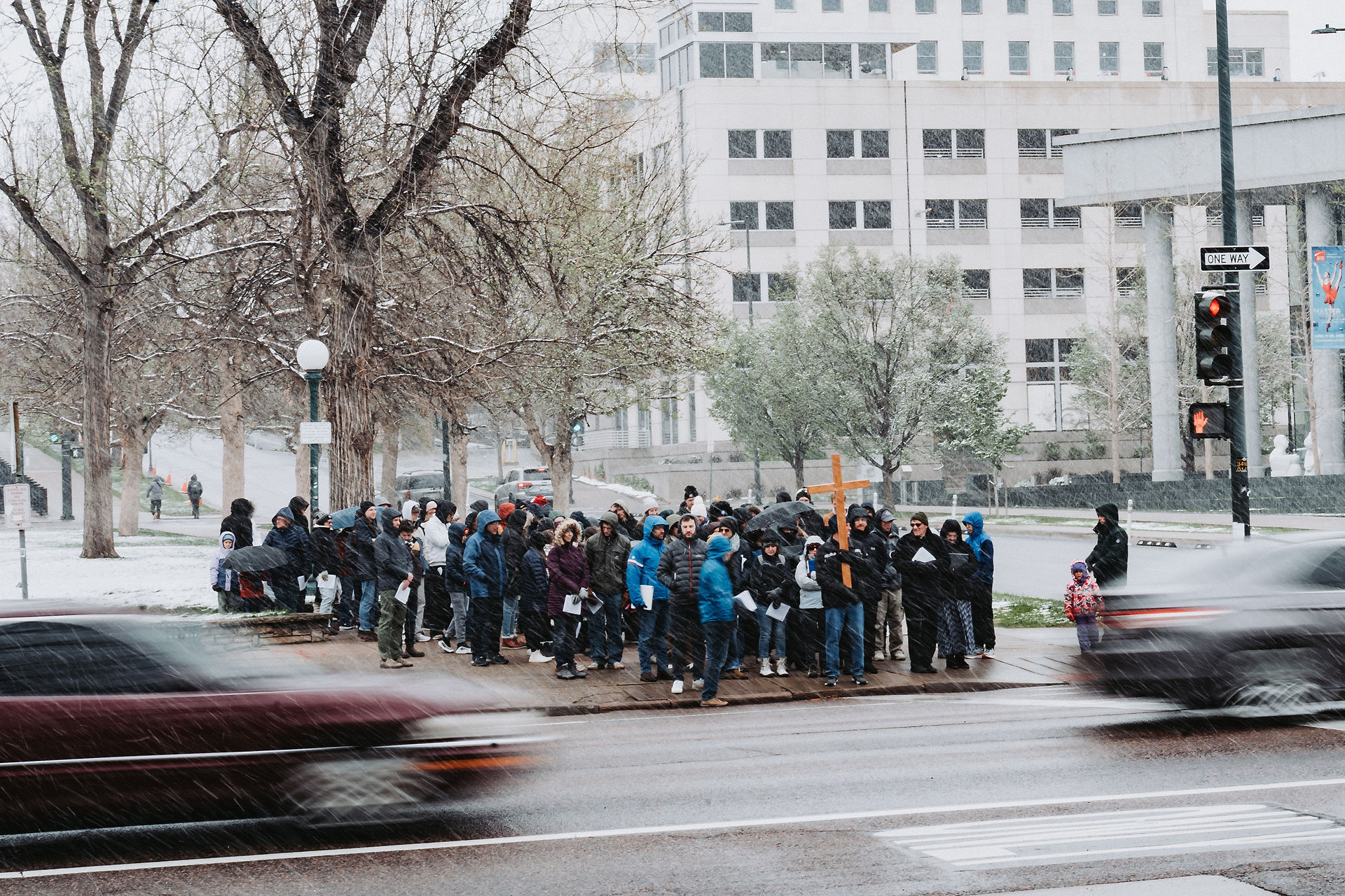 Crowd in winter coats stands on snowy sidewalk, holding a wooden cross. Cars, trees, and a building in the background; gray, overcast day.
