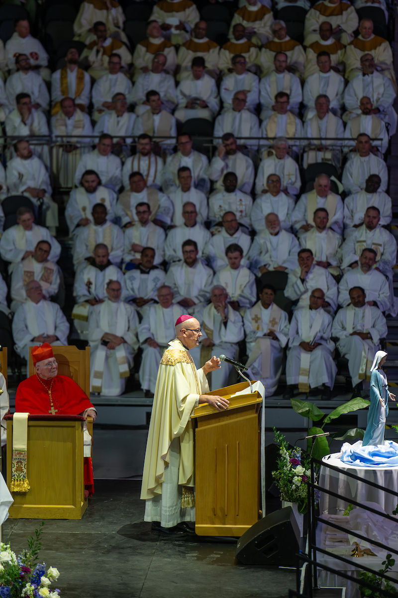 A bishop speaks at a wooden lectern, wearing liturgical vestments. A cardinal sits nearby. An audience of clergy is in the background.