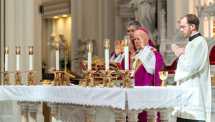 A religious ceremony with clergy in purple and white robes, praying at an altar with lit candles in an ornate church setting.