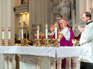 A religious ceremony with clergy in purple and white robes, praying at an altar with lit candles in an ornate church setting.