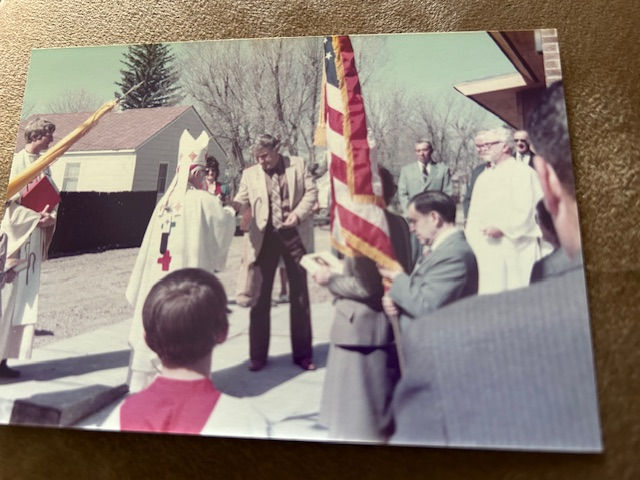 A group of people gather outdoors near a house, with one holding an American flag. Some are in robes, suggesting a formal or religious event.