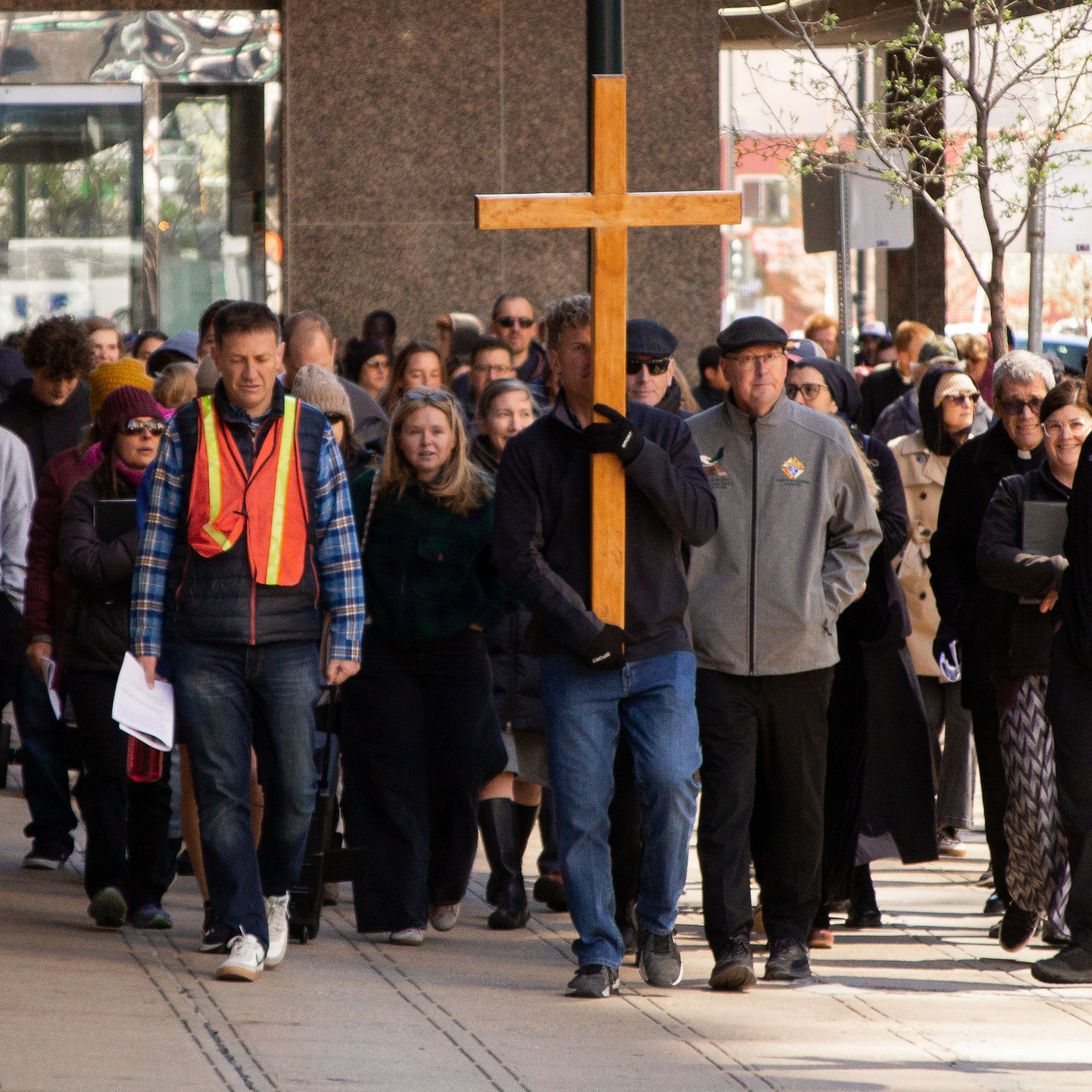 A large crowd follows a man carrying a wooden cross through a downtown sidewalk during a Way of the Cross procession.