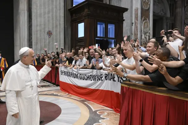 A religious leader greets a group of smiling people behind a red and white banner in a grand hall. The atmosphere is joyous.